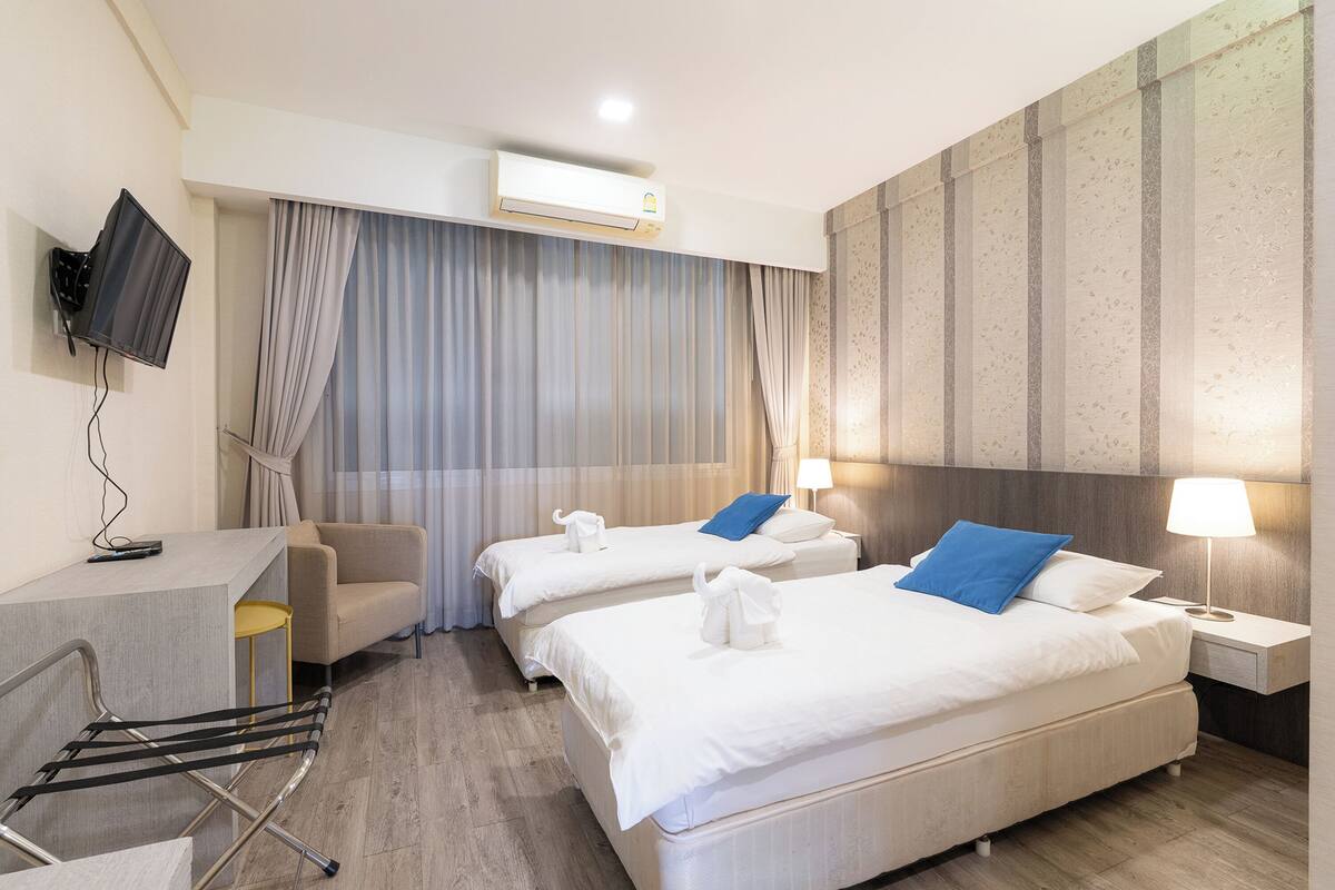 A hotel room with two beds, a desk, a chair, a TV, and an air conditioning unit.