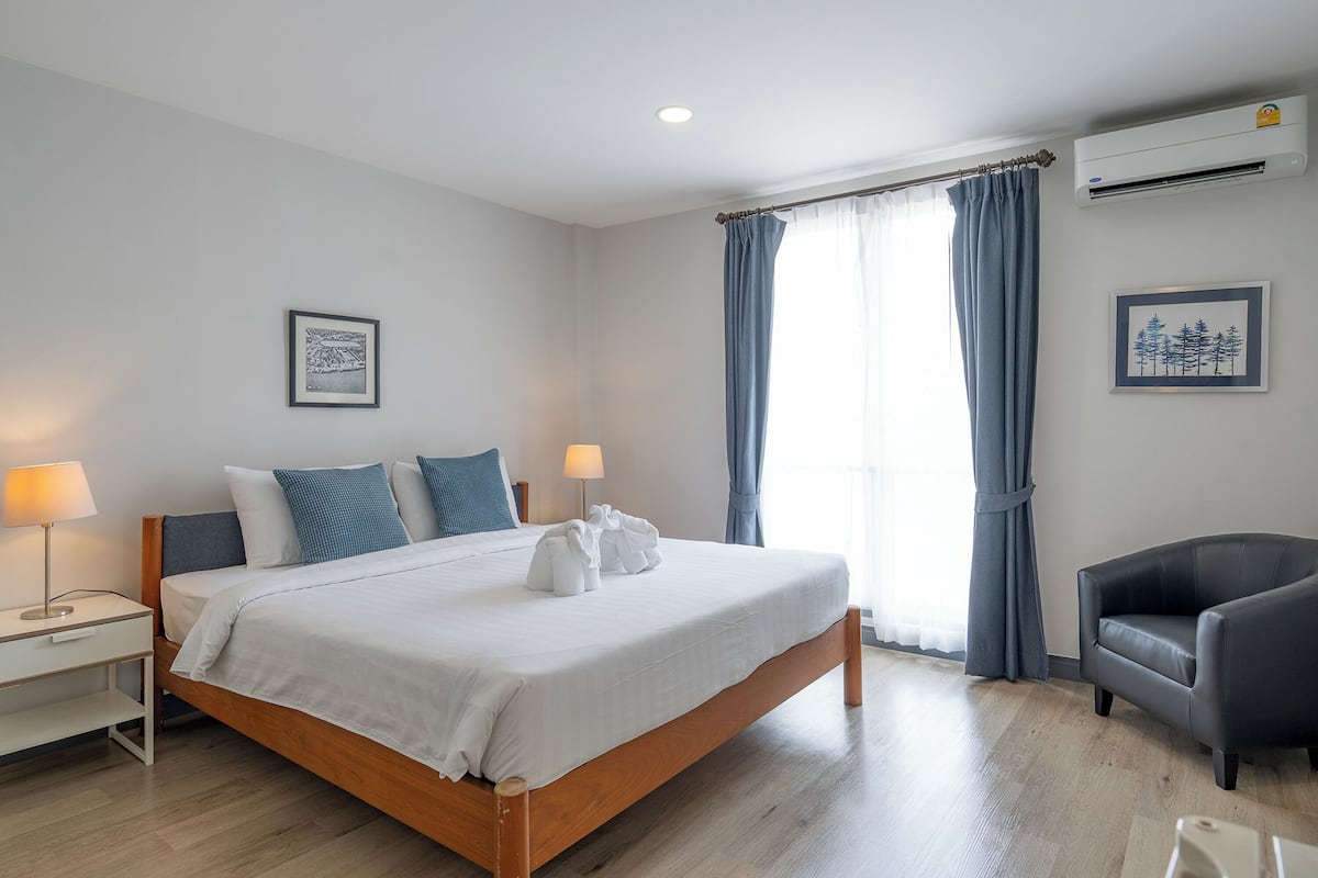 A hotel room with a wooden bed, two bedside lamps, a grey armchair, and a window with curtains.