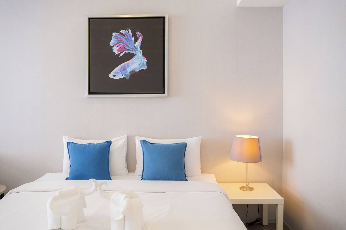 A hotel room with a bed, two blue pillows, a white bedspread, a framed artwork of a fish, and a bedside lamp.
