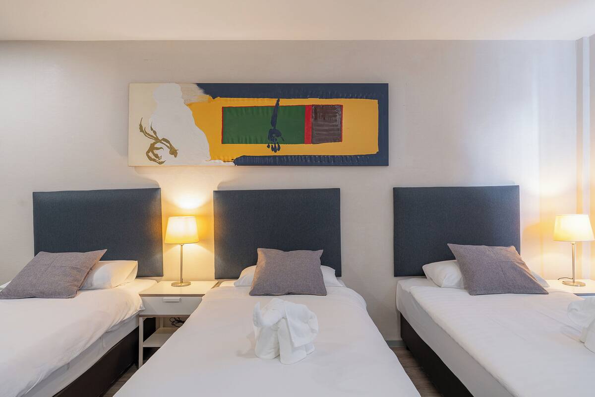 A hotel room with three single beds, each with a nightstand and lamp, and a large abstract painting above the beds.