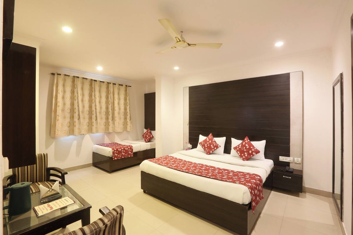 A hotel room with a large bed, a smaller bed, a desk with a chair, a ceiling fan, and a window with curtains.
