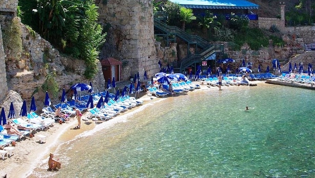 Een strand met helder water, blauwe parasols en ligbedden.