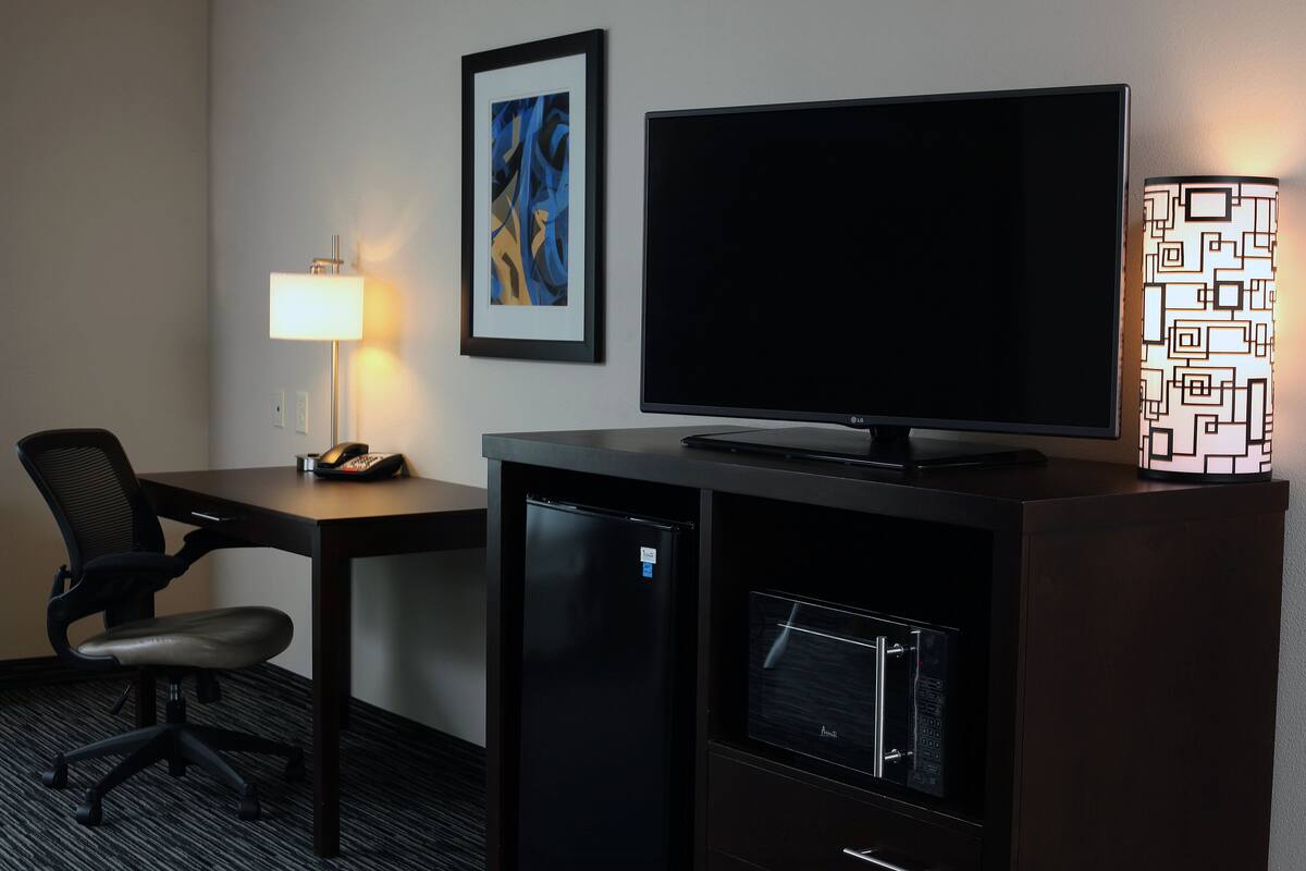 A hotel room with a desk, chair, television, refrigerator, and a lamp.