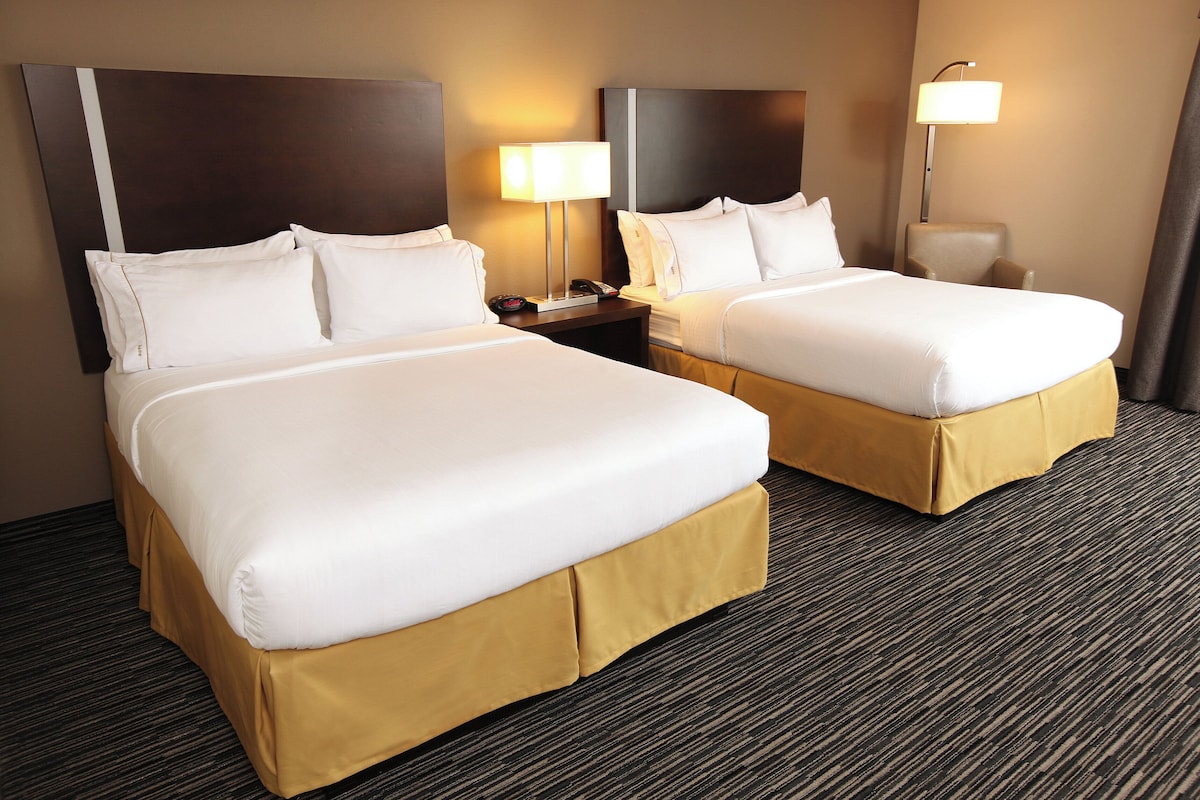 A hotel room with two beds, a nightstand with a lamp, and a chair.