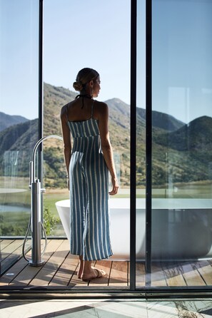 A woman in a striped dress standing by a glass-enclosed bathtub with a view of a mountainous landscape.