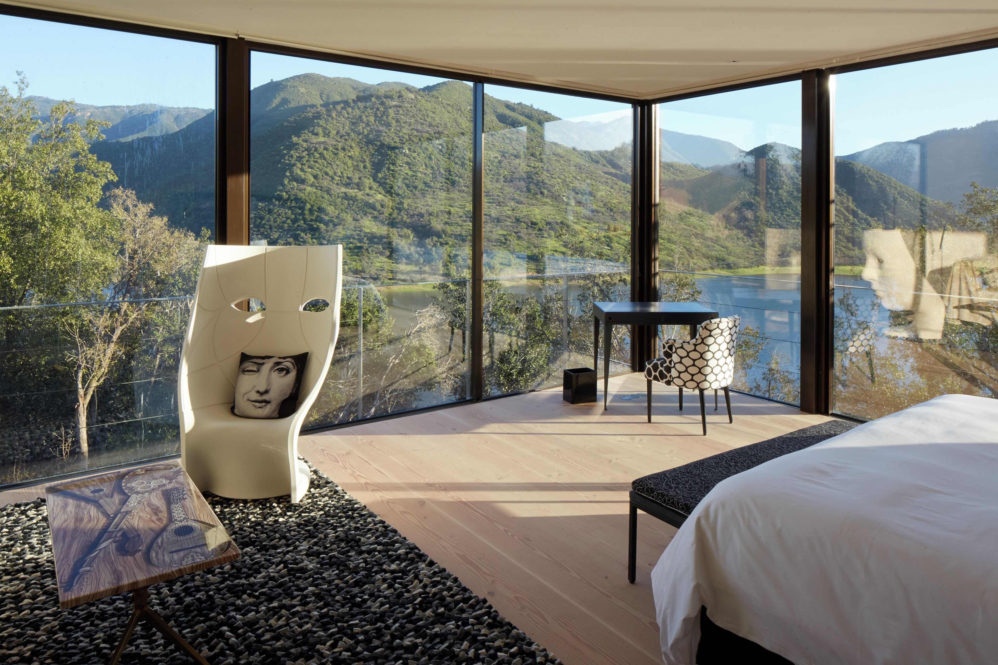 Un dormitorio moderno con una cama grande, una silla original con diseño de rostro, una mesita y vistas a un lago y montañas.