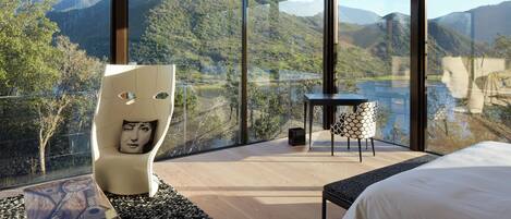 Un dormitorio moderno con una cama grande, una silla original con diseño de rostro, una mesita y vistas a un lago y montañas.