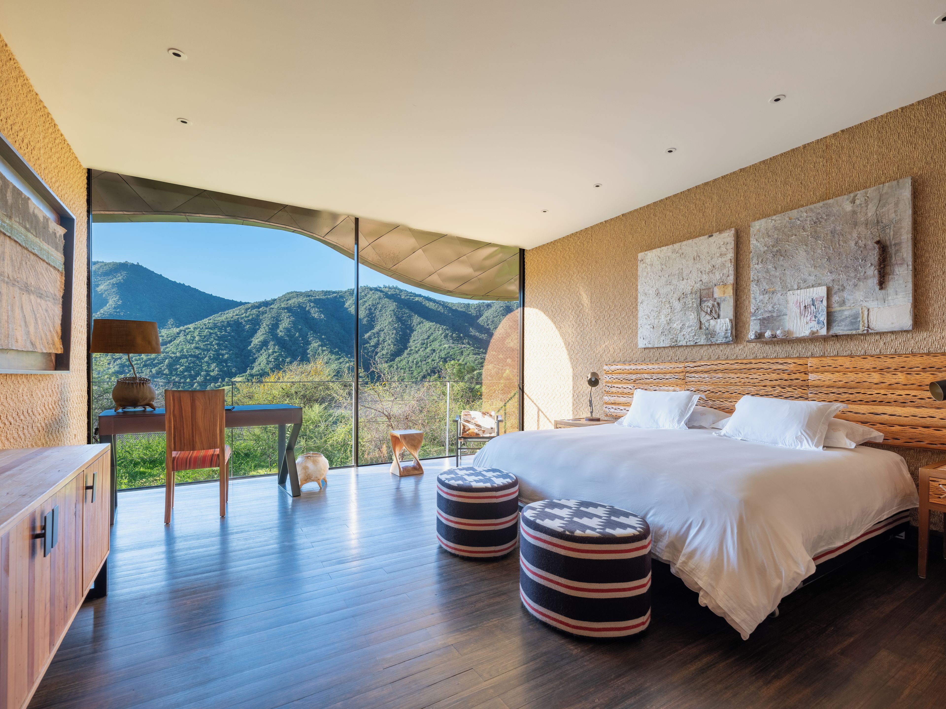 Quarto com uma cama grande, mesas de cabeceira, escrivaninha e uma cadeira. Vê-se montanhas através de uma janela ampla.