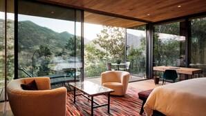 A modern bedroom with a large bed, a sofa, a small table, and a desk. The room has large windows offering a view of mountains and trees.