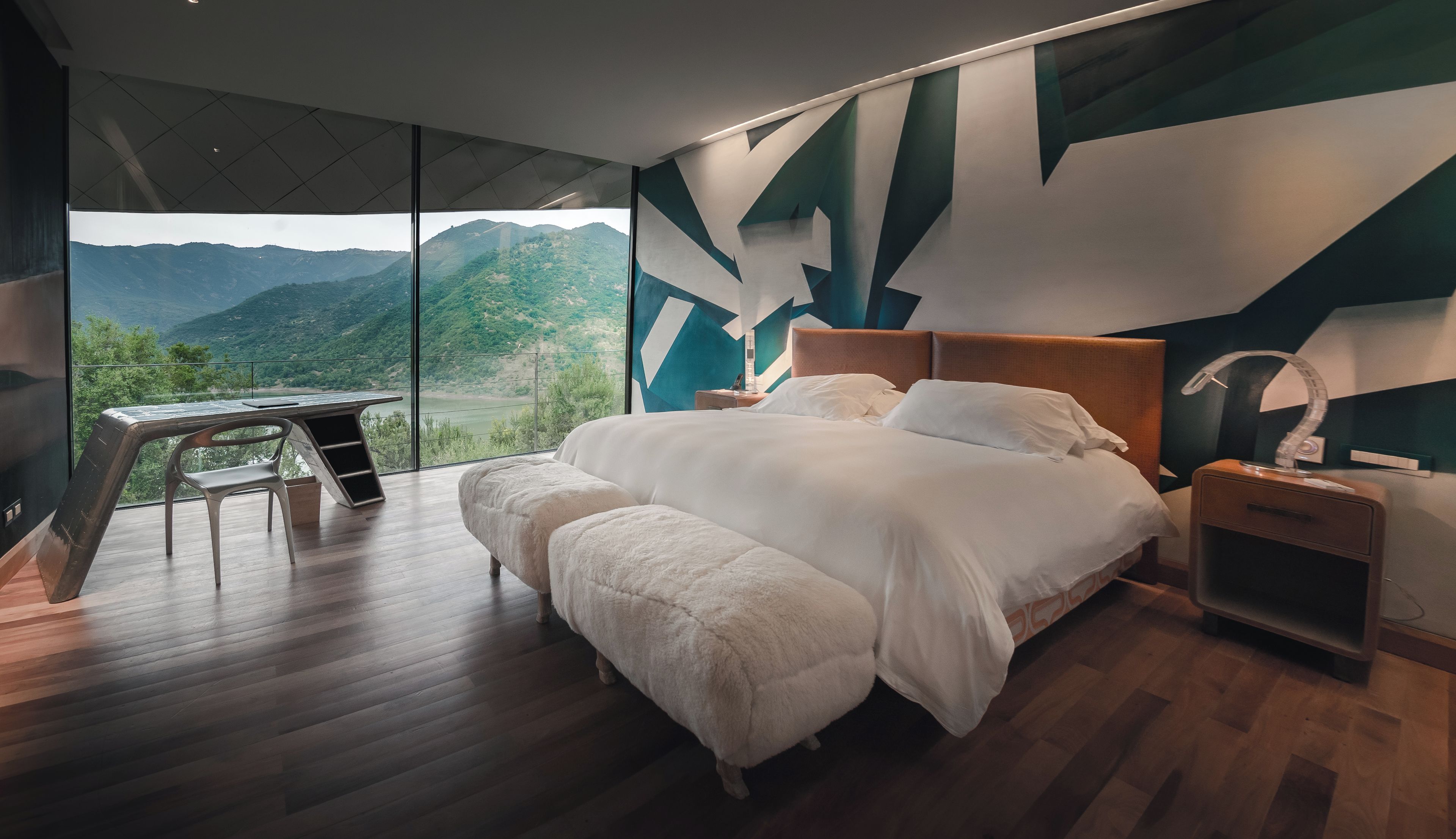 A modern bedroom with a large bed, a desk, and a view of mountains through a large window.