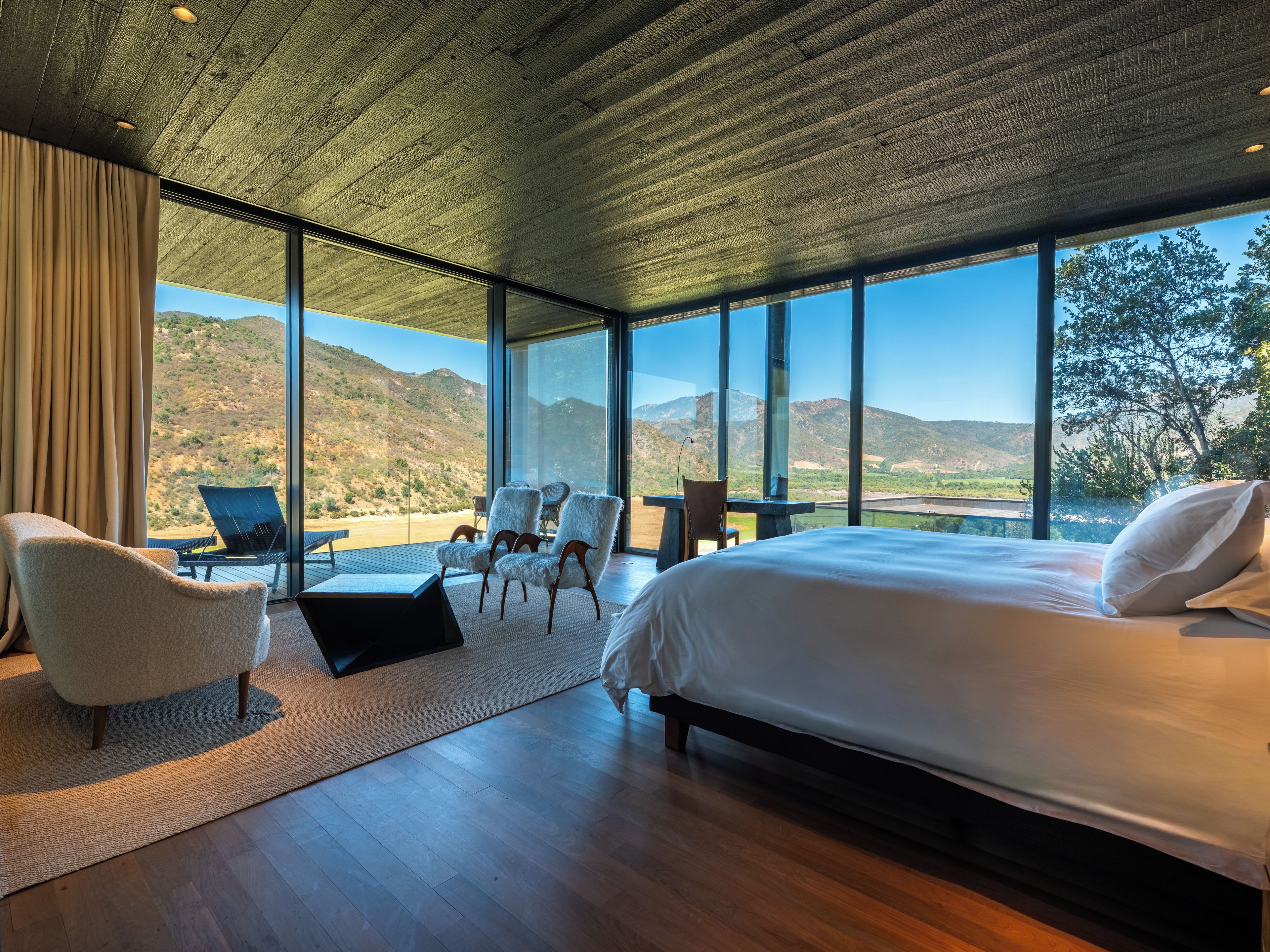 A modern bedroom with a large bed, two chairs, a small table, and a view of a mountainous landscape through large windows.