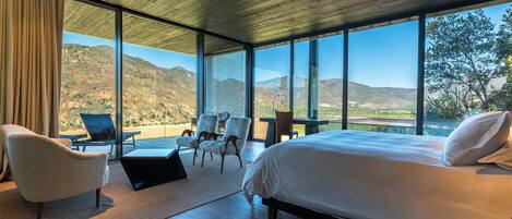 A modern bedroom with a large bed, two chairs, a small table, and a view of a mountainous landscape through large windows.