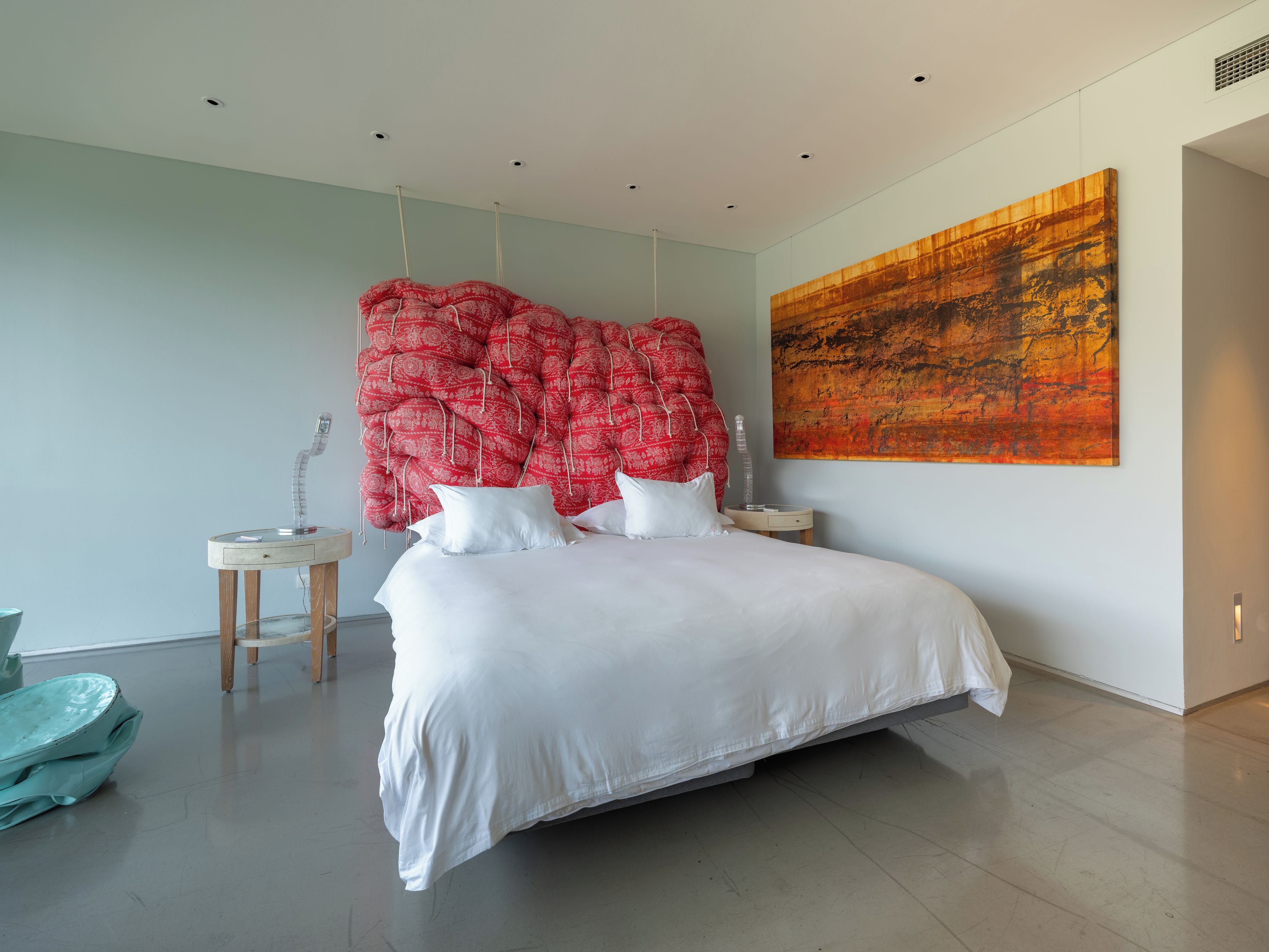 A modern bedroom with a large bed, a unique red headboard, a small bedside table, and a large abstract painting on the wall.