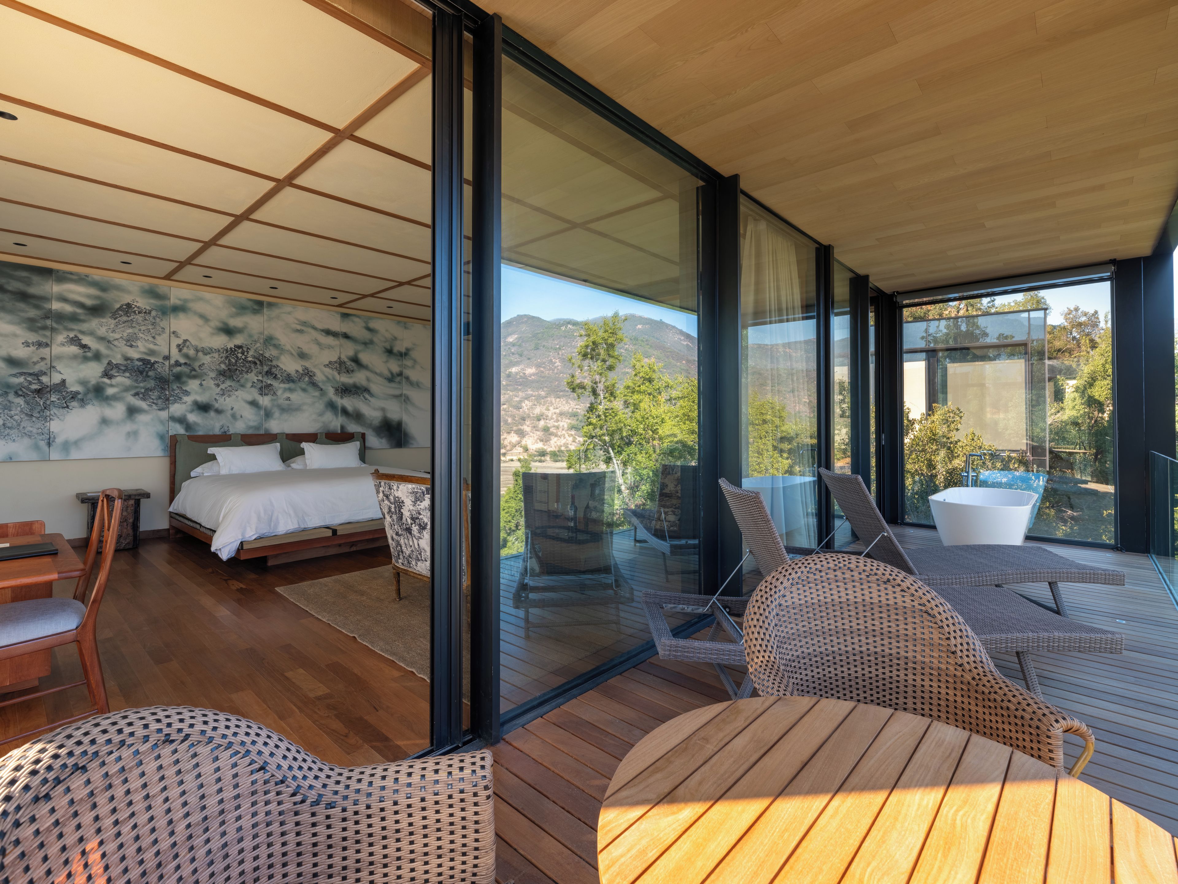 A modern bedroom with a large bed, a wooden floor, and a view of the outdoors through large windows.