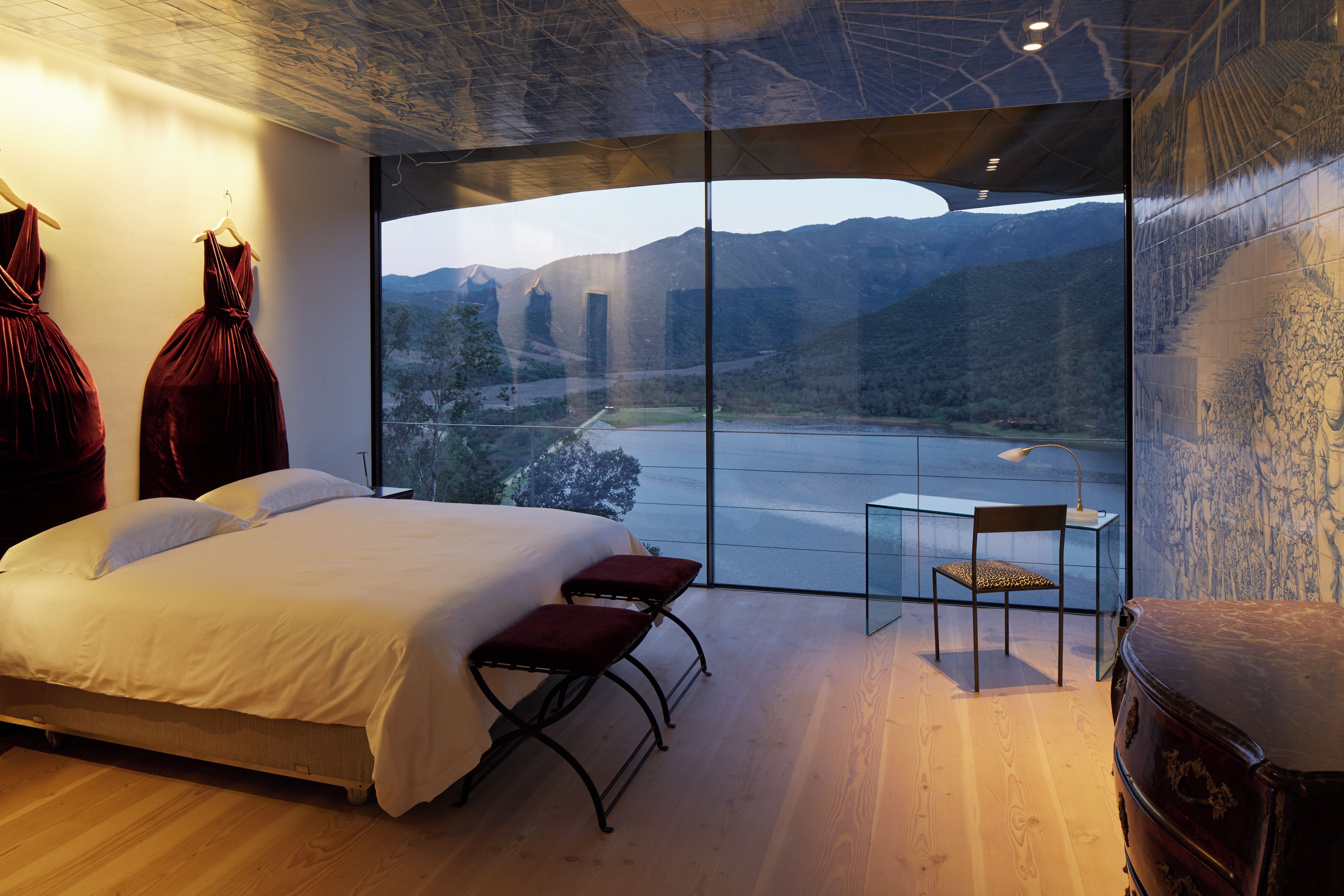 Un dormitorio con una cama grande, una mesita pequeña y una silla, con vistas a montañas y un río a través de ventanales de piso a techo.