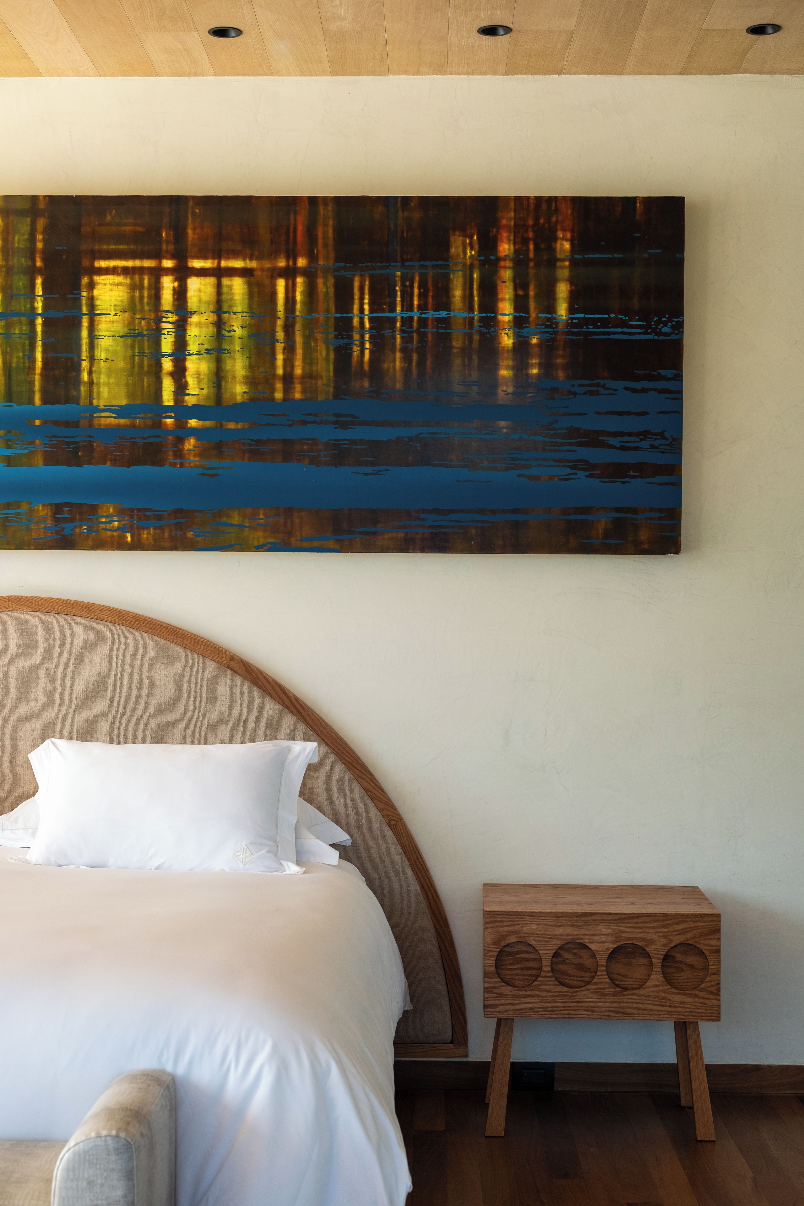 A bedroom with a bed, a headboard, a wooden bedside table, and a large painting on the wall.