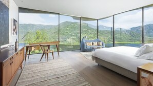 A bedroom with a large bed, a desk, and a chair, with a view of mountains and a lake.
