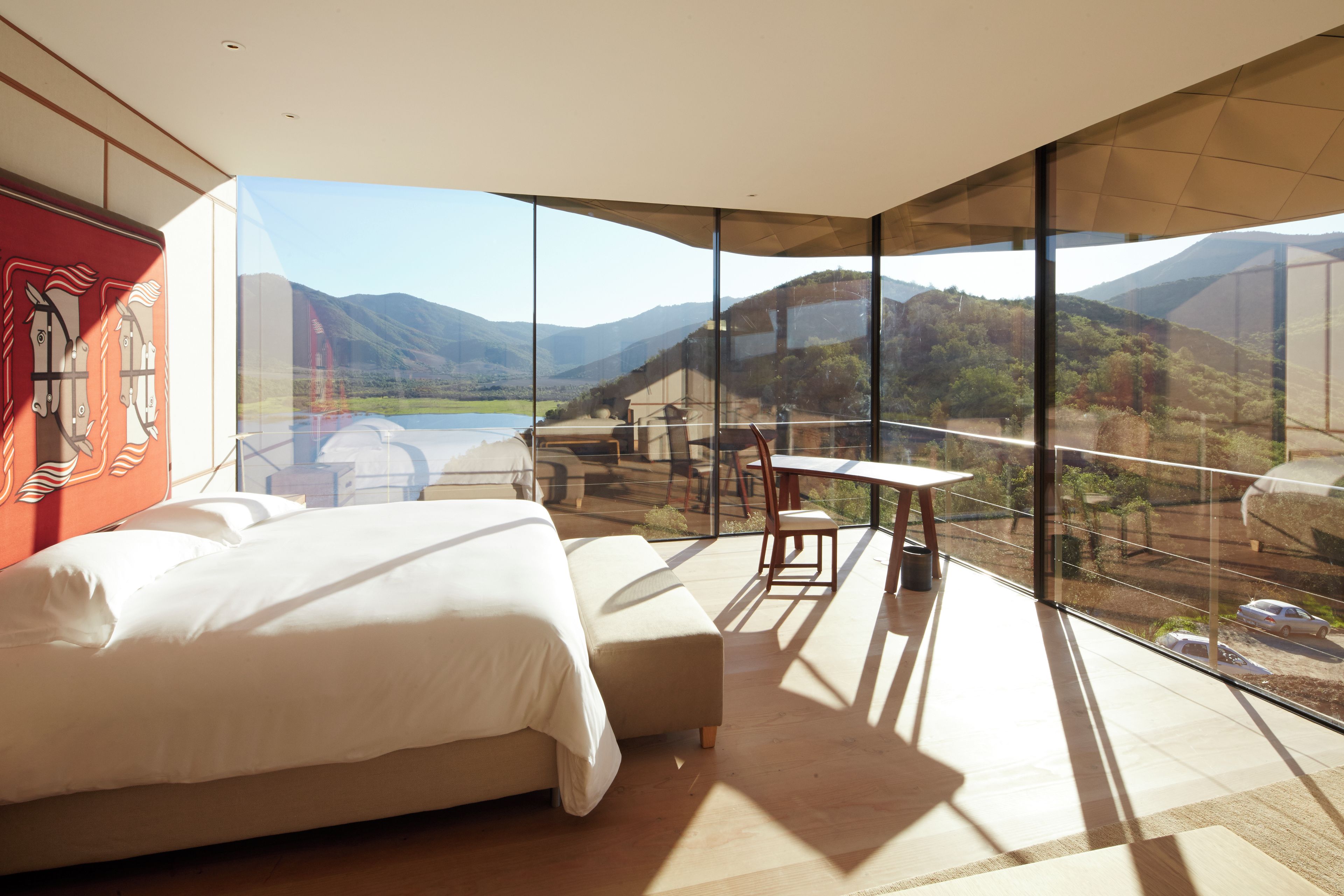 Un dormitorio moderno con una cama grande, una mesita pequeña y una silla. Cuenta con un amplio ventanal con vistas a la montaña.