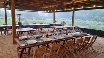 Un área de comedor al aire libre con mesas dispuestas para una comida, rodeada de sillas de madera y grandes ventanales con vistas a la vegetación.
