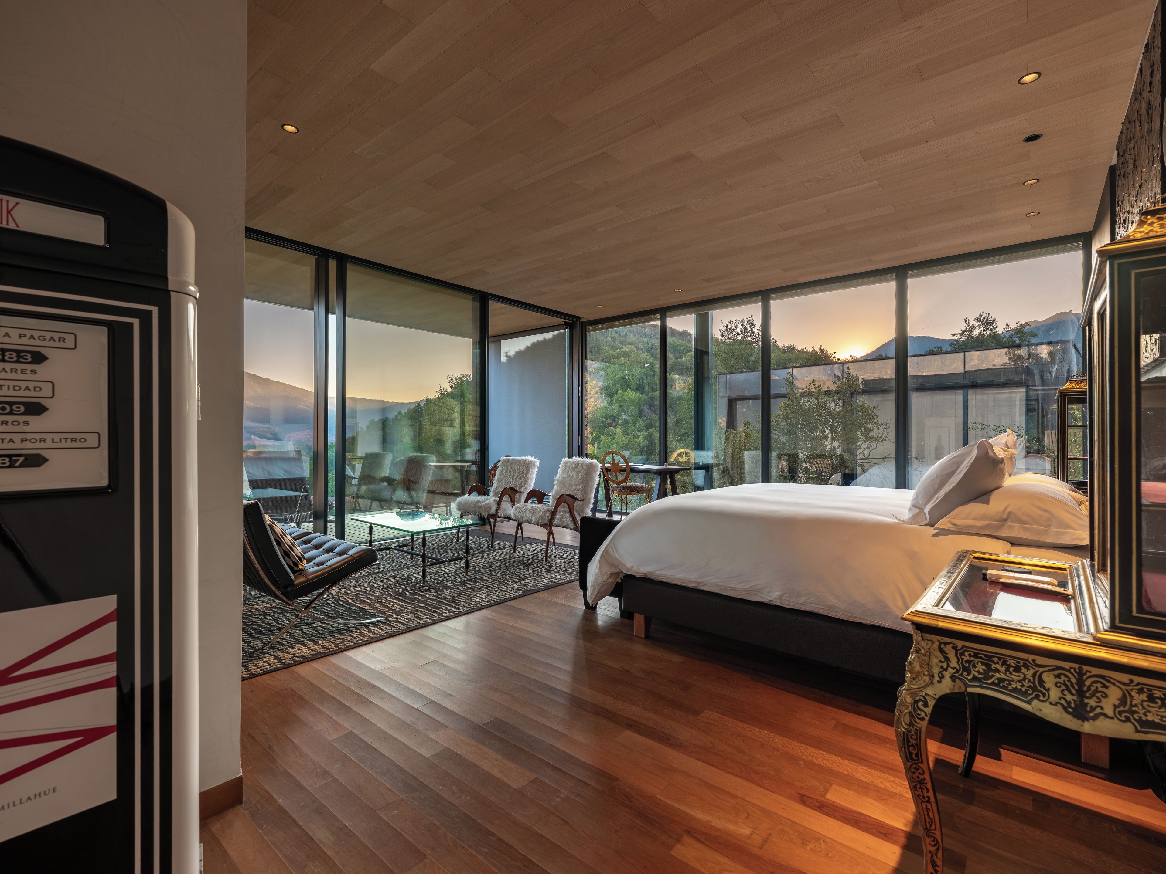 A modern hotel room with a large bed, a glass wall, and a view of the outdoors.