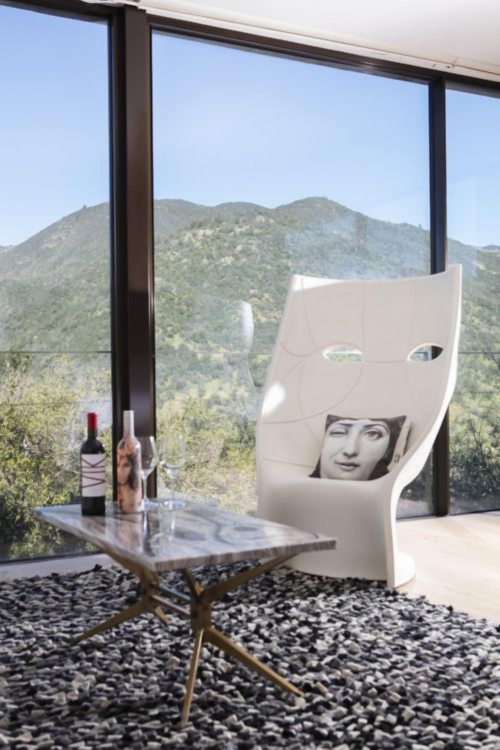 Una habitación moderna con un ventanal grande, una silla blanca con almohada para el cuello, una mesa de vidrio con botellas y un suelo de piedritas.