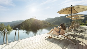 A wooden deck with a pool, lounge chairs, and umbrellas overlooking a lake with mountains in the background.