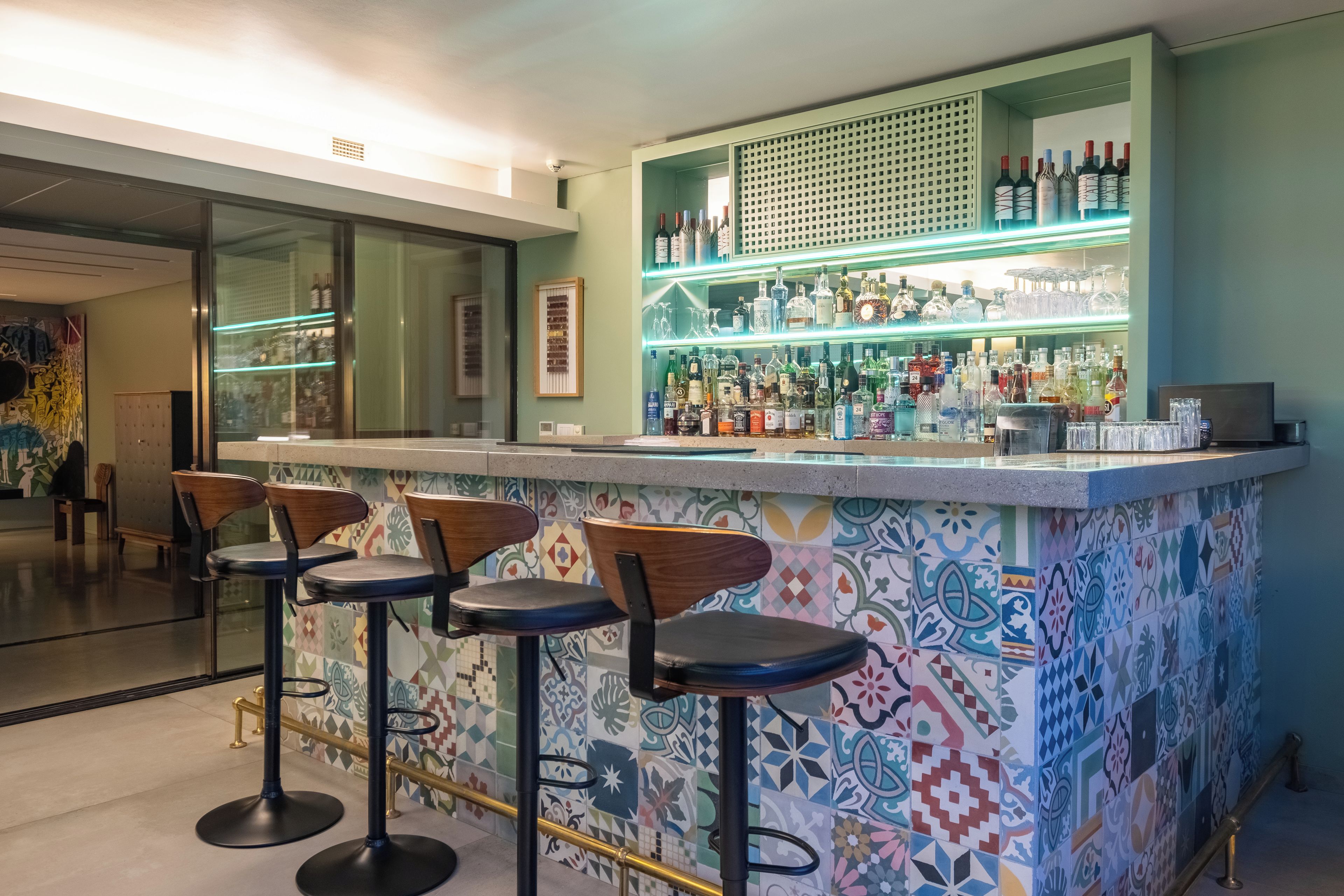 A bar area with a colorful tiled backsplash, bar stools, and a variety of bottles and glasses on shelves.