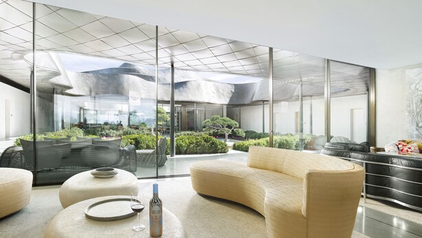A modern interior with a glass wall, a seating area with a sofa and ottoman, and a bottle of wine on a table.