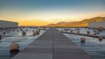 A pathway leading through a water feature with large rocks, surrounded by concrete walls and a hilly landscape in the background.