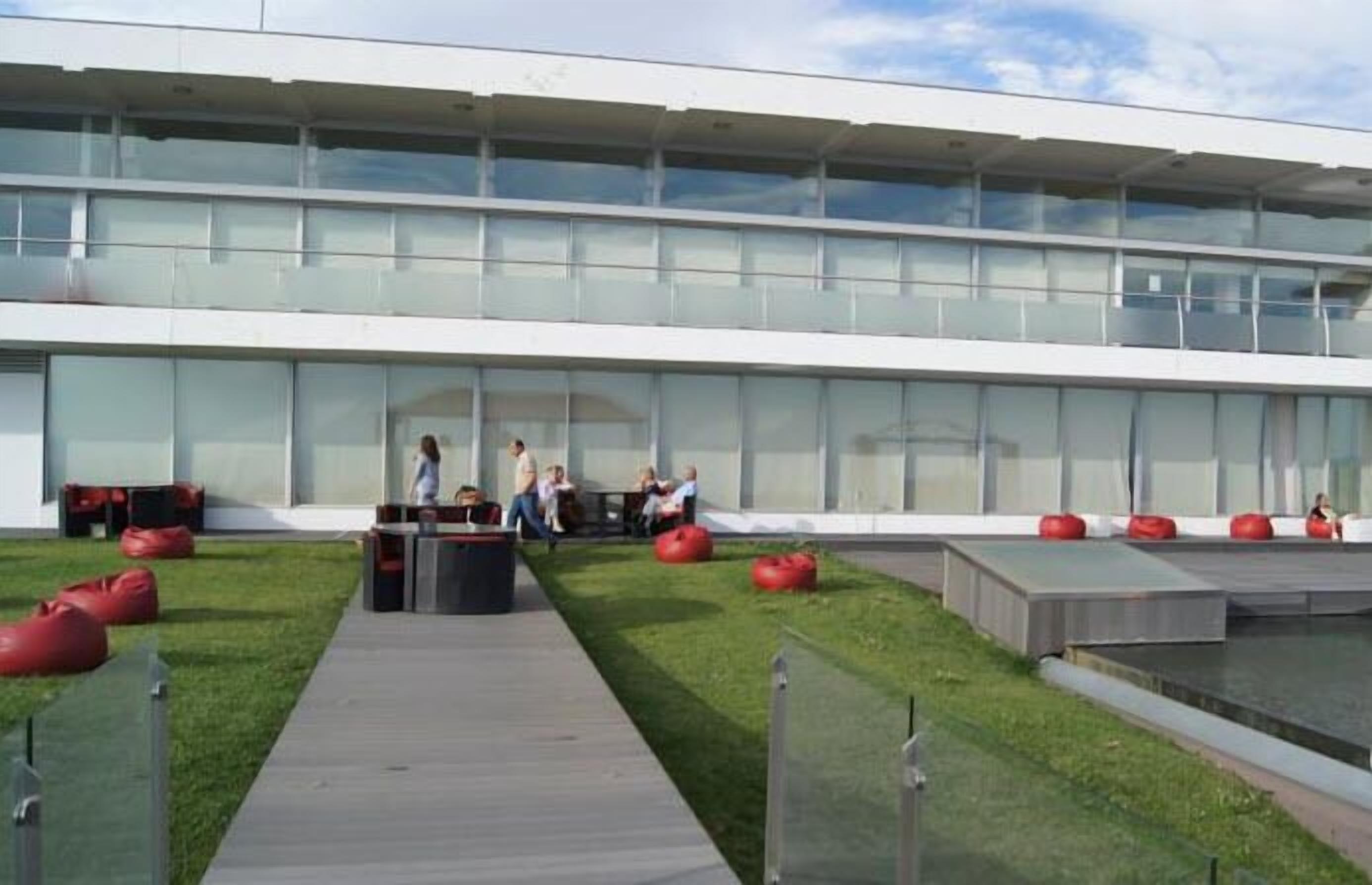 A modern building with a glass facade, outdoor seating area with red bean bags, and a pathway leading to the entrance.