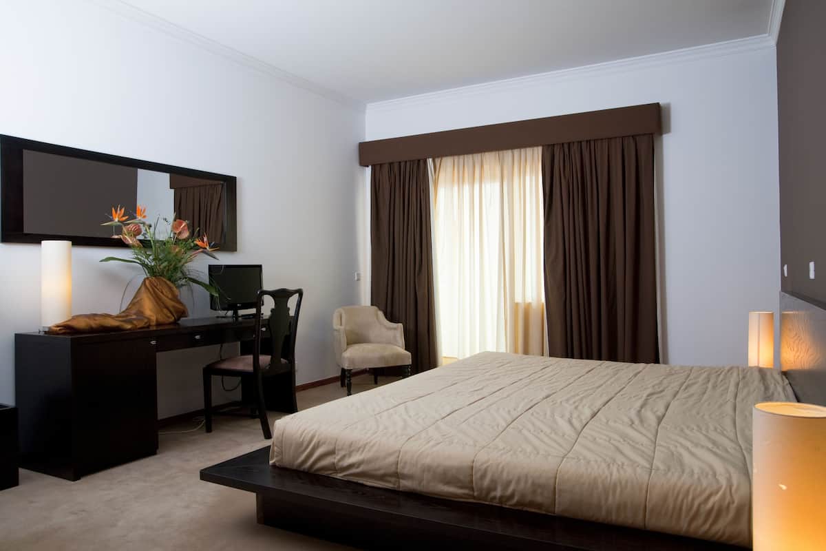 Une chambre d’hôtel comprenant un lit, un bureau, une chaise, une lampe de bureau, un miroir et une fenêtre avec des rideaux.