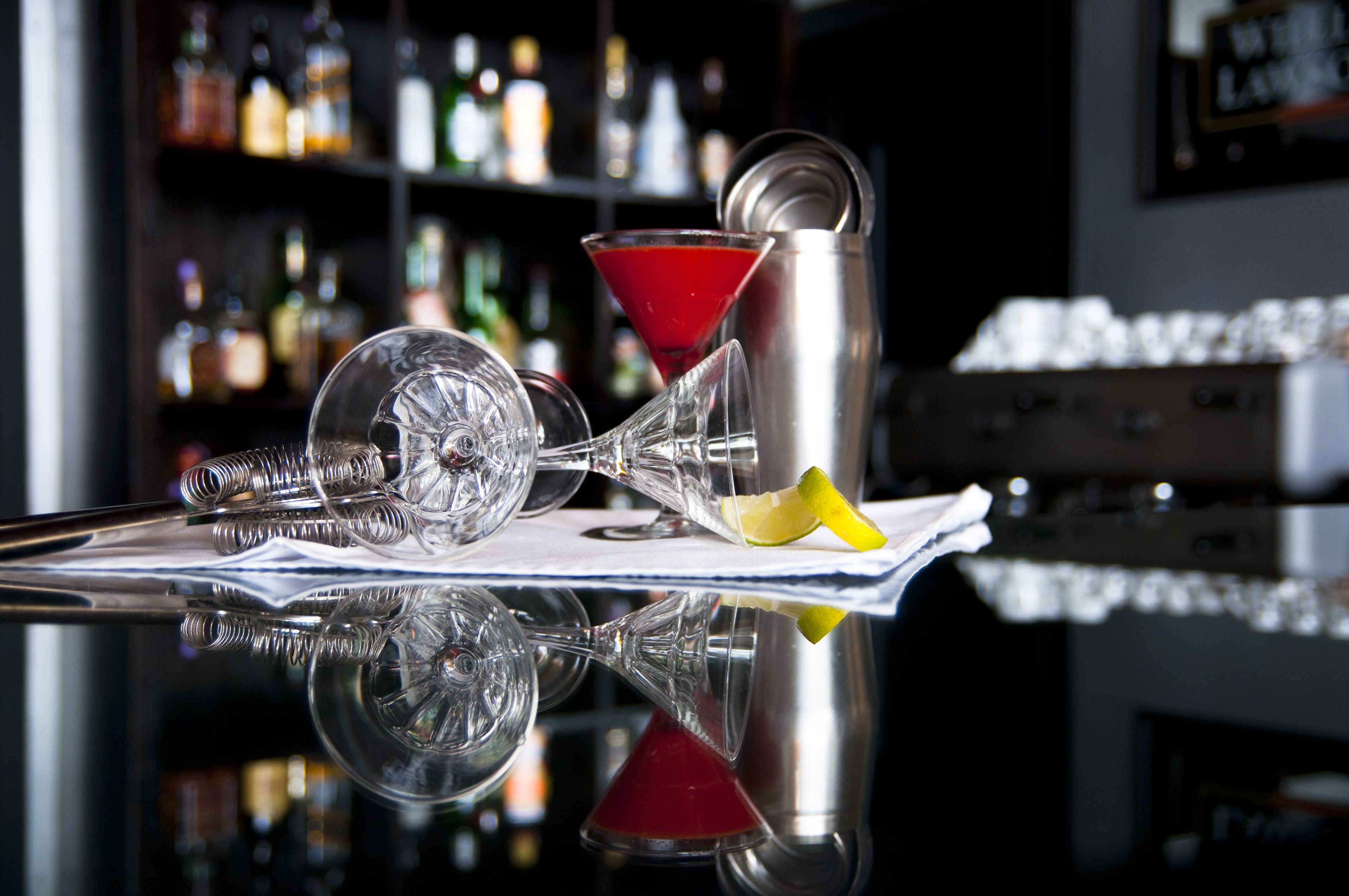 A bar setting with a martini glass, a cocktail shaker, and a bottle of liquor on a reflective surface.