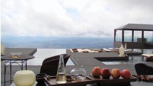 Ein Essbereich im Freien mit Blick auf den Ozean und die Berge, mit einem Tisch, der mit Früchten, einer Vase und einer Flasche dekoriert ist.