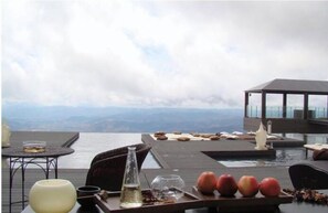 An outdoor dining area with a view of the ocean and mountains, featuring a table with fruits, a vase, and a bottle.