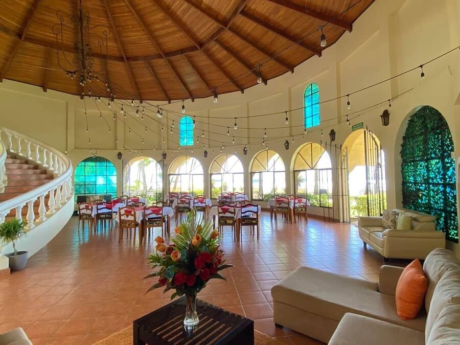 Un espacio interior amplio con techo de madera alto, varios ventanales arqueados y una zona de estar central con mesa y sillas.