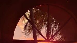 A sunset view through a large arched window with palm trees and a table visible inside. - Hotel Delfin Playa Bejuco (Parrita)