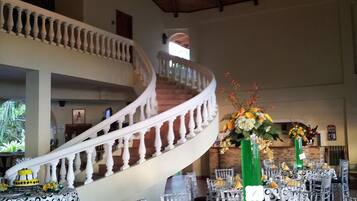 A spacious hall with a grand staircase, clear glass chairs, and tables set for a formal event.