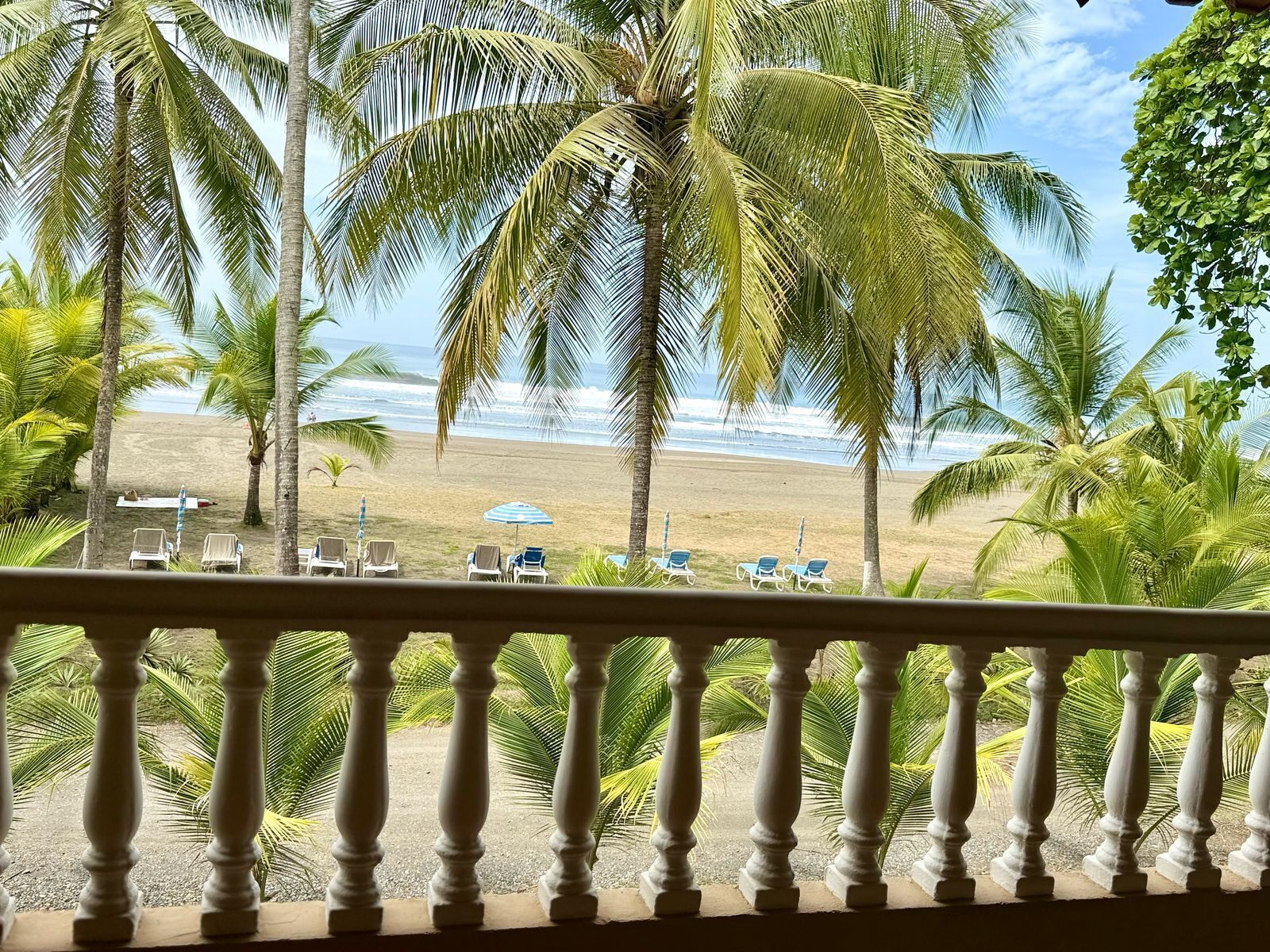 A balcony with a view of a beach, palm trees, and lounge chairs.