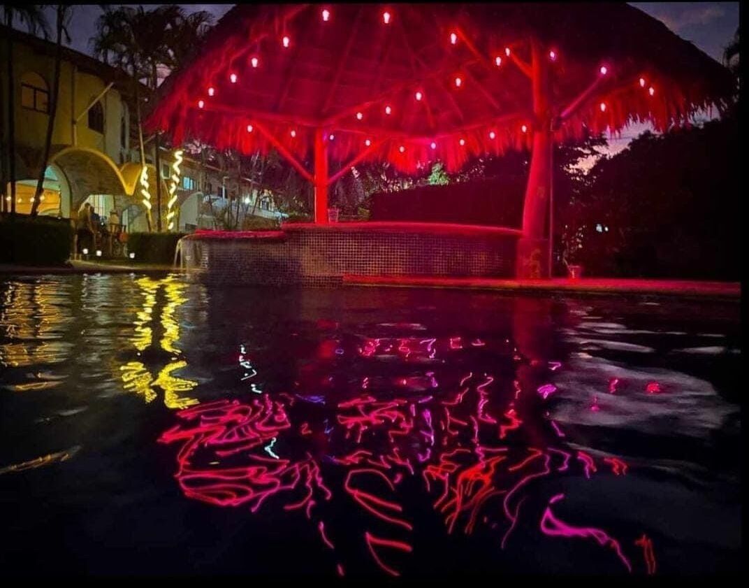 A nighttime scene with a water feature reflecting red lights, a covered seating area, and a building in the background.