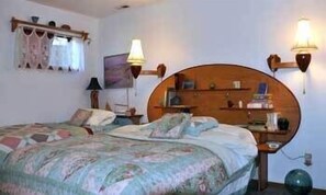 A bedroom with a wooden headboard, two bedside tables, a lamp, a shelf with books, and a window with curtains.