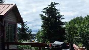 A wooden deck with a table and chairs, a grill, and a building in the background. - Whalesong Bed and Breakfast (Homer)
