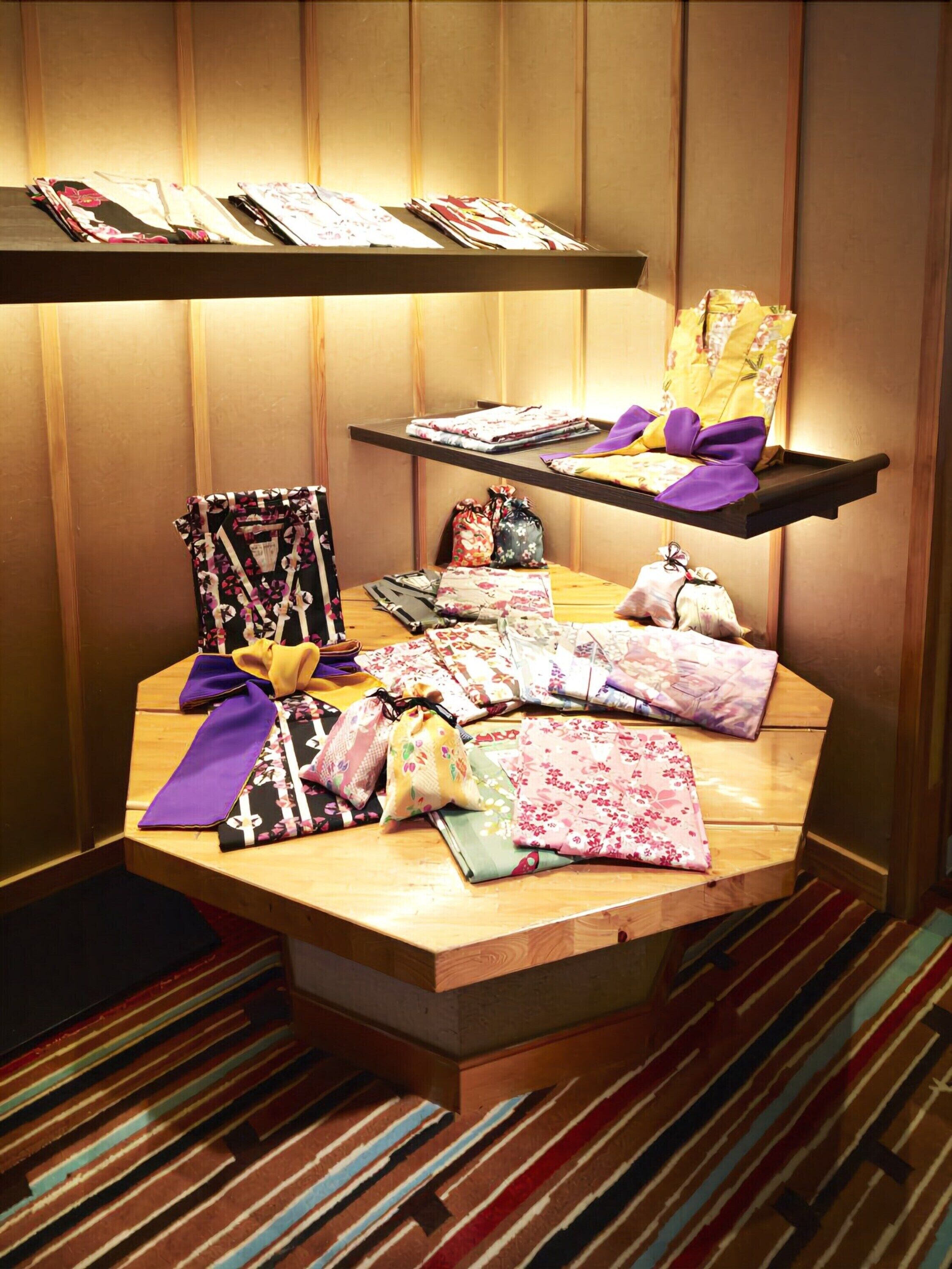 Una exhibición de varias telas y textiles sobre estantes de madera y una mesa, con una alfombra a rayas visible en primer plano.