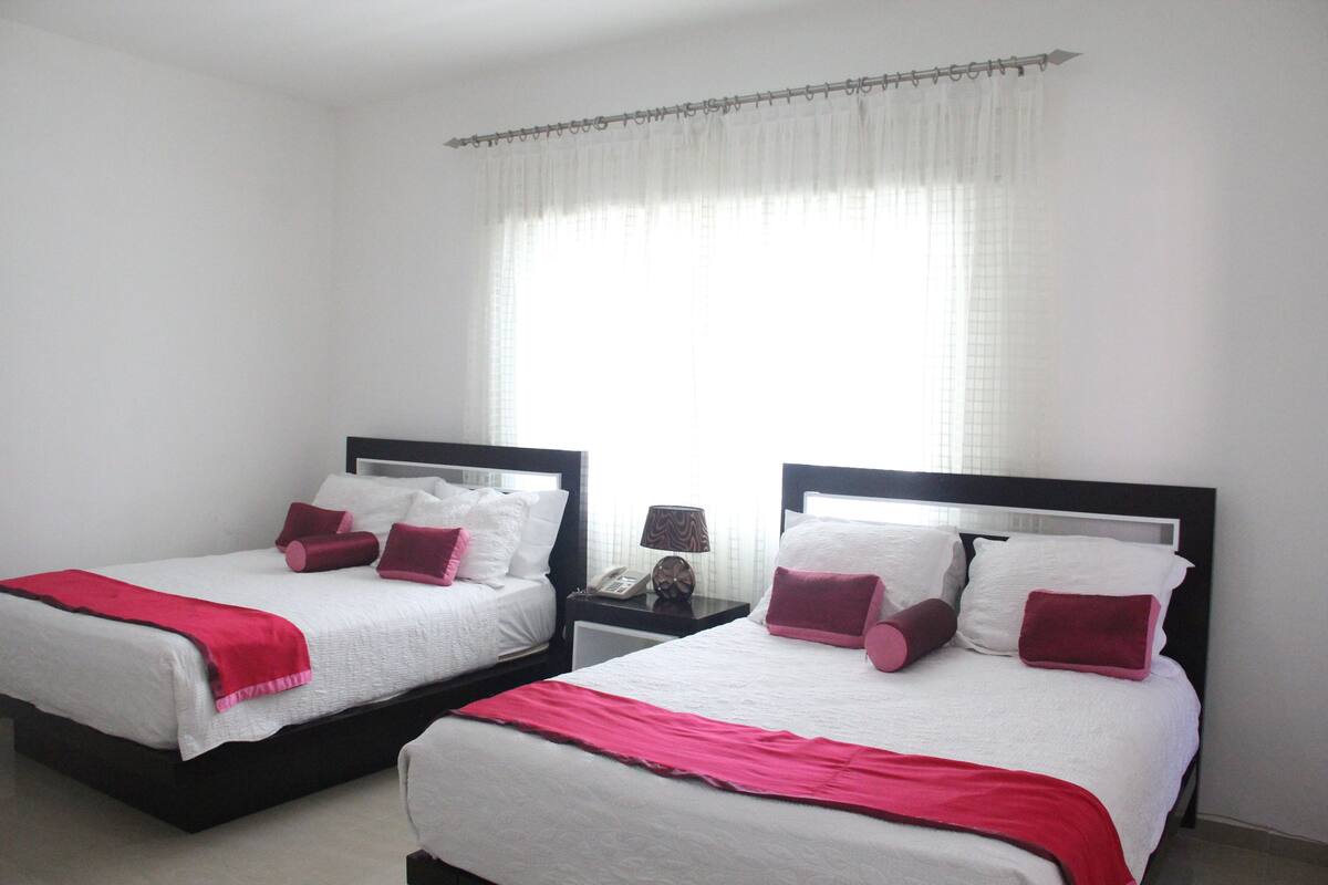 Une chambre d’hôtel avec deux lits, chacun agrémenté de draps blancs et de touches de couleur rouge, une table de chevet avec une lampe et une fenêtre ornée de rideaux légers.
