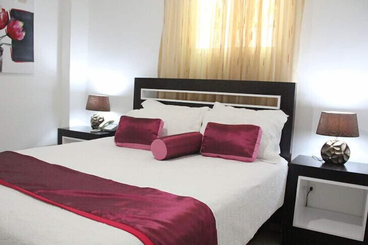 Un lit bien fait, recouvert d’une couvre-lit rouge et blanche, avec deux oreillers rouges et un plaid assorti. Il y a deux tables de chevet avec des lampes, et l’une d’elles est ornée d’un objet décoratif.