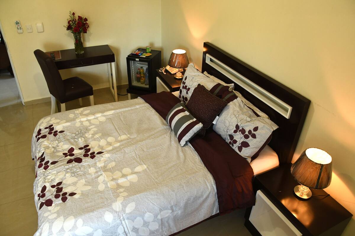 Un lit bien fait, recouvert d’une couvre-lit à motifs floraux, une lampe de chevet et une petite table de nuit avec une lampe et un vase de fleurs.