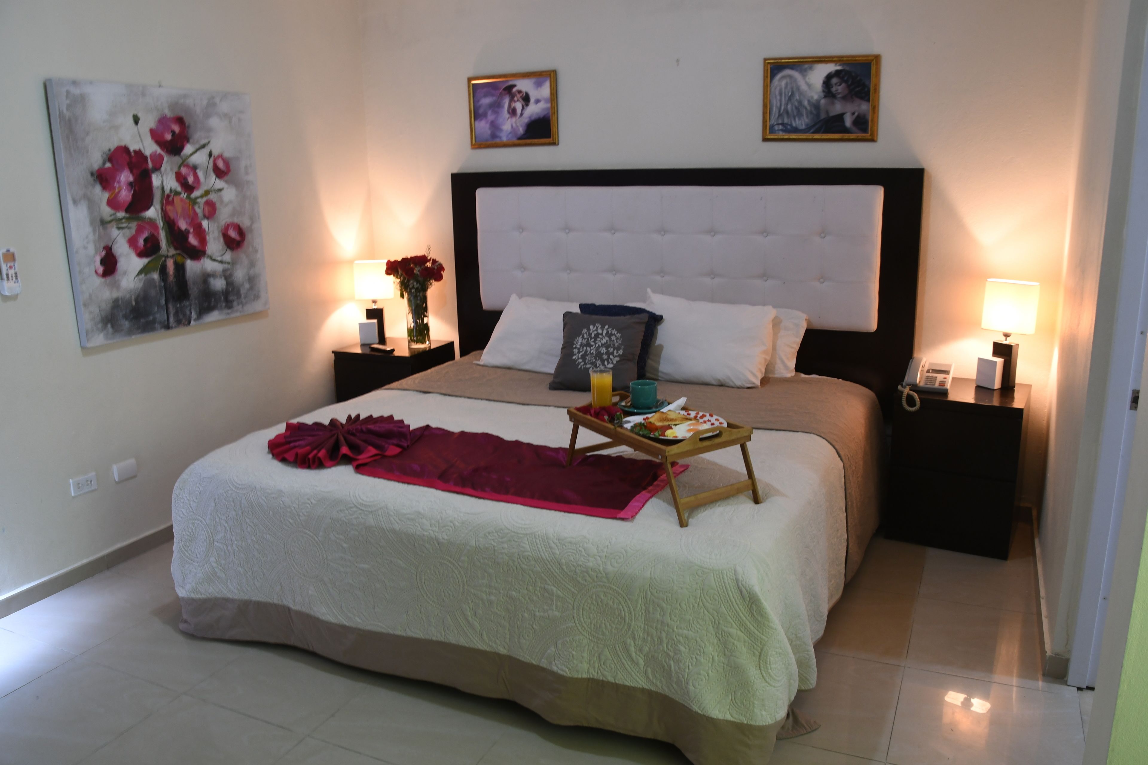 Una cama bien hecha con un plaid rojo, una mesita de noche con una lámpara y un cuadro en la pared.