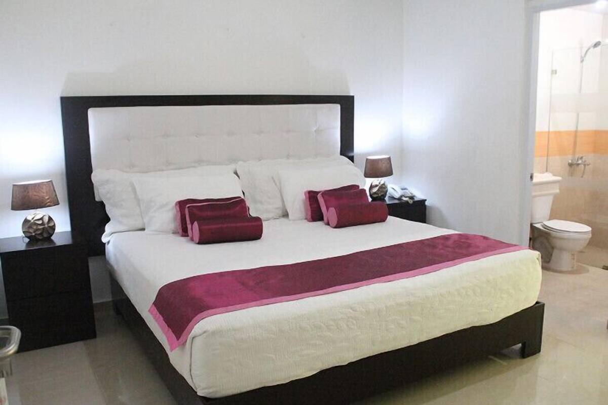 Un lit bien fait, recouvert d  ’une couvre-lit blanche et rose, deux tables de chevet avec des lampes, et une salle de bain visible au fond.