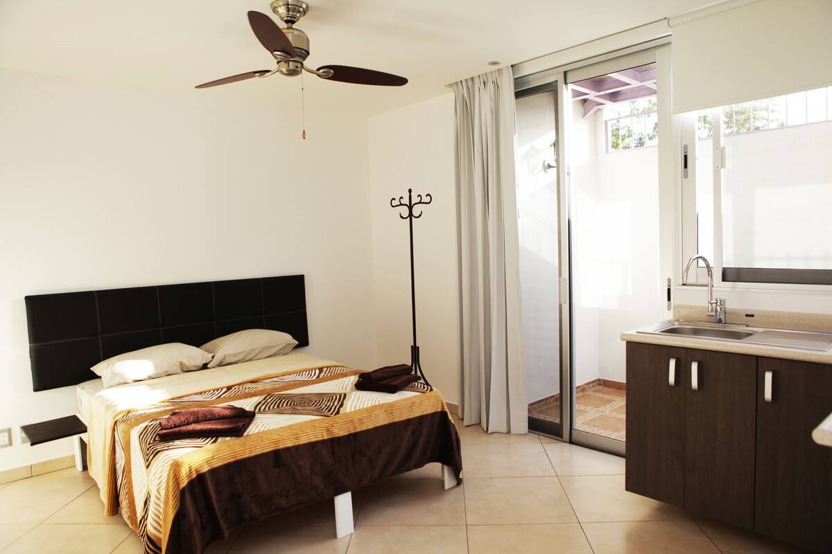 A bedroom with a bed, a ceiling fan, a coat rack, a bathroom with a sink, and a sliding glass door to a balcony.
