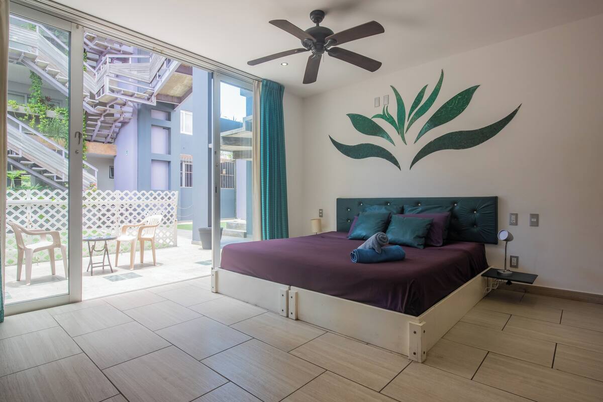 A bedroom with a bed, a ceiling fan, a wall mural, a sliding glass door leading to an outdoor area, and a small table with a lamp.