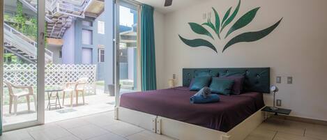 Une chambre à coucher comprenant un lit, un ventilateur de plafond, une fresque murale, une porte-fenêtre coulissante donnant sur un espace extérieur, et une petite table avec une lampe.